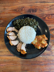 Savory fried pork belly slices accompany a mound of rice and sautéed greens, presented on a black plate atop a rustic wooden table