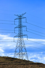 Photograph of a large electricity Transmission Tower on a grassy hill against a bright blue sky in...