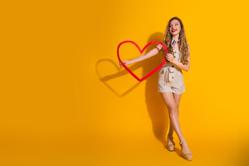 Smiling young woman holding red heart frame in yellow background embodies happiness, charm, and...