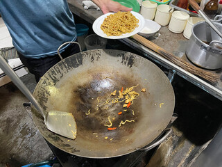 A cook is stirring noodles in a large wok while serving a plate of freshly fried noodles in a bustling kitchen