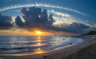 Panorama of a resort beach ad sea at sunset. The sky is covered with clouds above the sun. Beautiful reflections of the sunset on the water. Mediterranean, Side,  Antalya province, Turkey