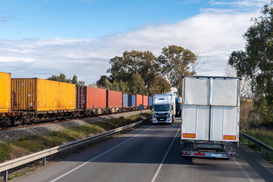 Trucks driving on a two-way road next to railway tracks with a train carrying intermodal container wagons.