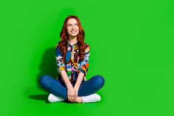 Stylish young woman with red hair sitting against a green background, wearing colorful casual...