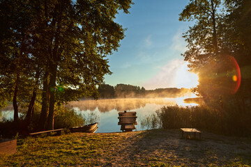 A peaceful lakeside scene at sunrise with mist rising from the water, surrounded by trees. A wooden...