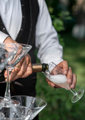 hands of the barman, in white shirt and black vest, pouring champagne from a bottle of 