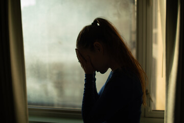 silhouette of a woman next to apartment window hand over face feeling depressed, sadness,...