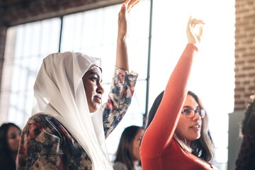 Woman in hijab and diverse group of people raising hands in a Seminar room setting, showing...