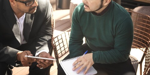Two men in discussion, one holding a tablet, the other a notebook. Engaged in conversation, sharing ideas, collaborating. Professional setting, focused interaction. Men having conversation in cafe.