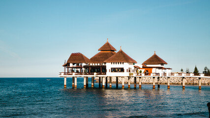 An elegant stilted structure with thatched roofs extends over the blue waters of M'diq, surrounded by a wooden walkway and greenery.