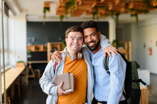 Young man with Down syndrome and his buddy with arms around looking at camera indoors at school