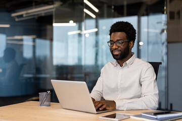 Young successful businessman working on laptop inside office, man in light shirt smiling contentedly, typing on laptop keyboard at workplace.