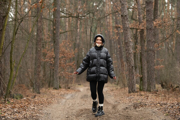 In a serene forest setting, a young woman walks joyfully along a dirt path. She wears a cozy black puffer jacket, black leggings, and cheerful boots, enjoying the cool autumn weather