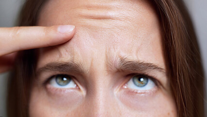 Frown lines. Botox injections. Botulinum toxin. Cropped portrait of a woman with light skin and blue-green eyes examining forehead wrinkles with a concerned expression. .