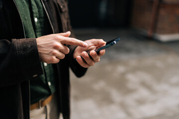 Cropped shot of unrecognizable man holding and using smartphone on city street, browsing internet, checking social media, using modern mobile app. Concept of modern communication and urban lifestyle.