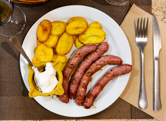 Fried butifarra sausages with fried potato slices are served on plate. Food is complemented by bowl with spicy white garlic alioli sauce