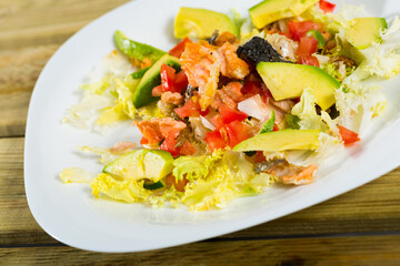 Salad of grilled salmon, fresh avocado, lettuce, tomatoes and lemon served on white platter on wooden background