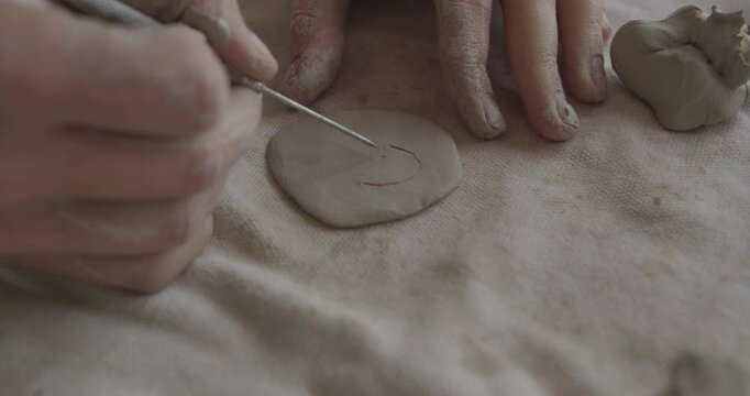 Hand engraves a circular pattern on a flat clay surface using a sculpting tool. The creative process highlights fine detail work and craftsmanship in handmade ceramic art.