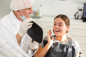 Elderly male dentist showing young female patient result of dental procedure in dental clinic