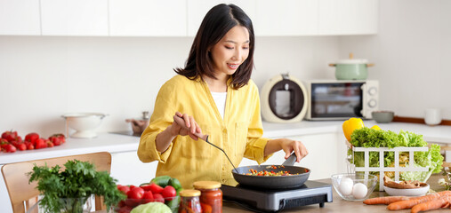 Young Asian woman cooking different vegetables in frying pan on portable electric stove at kitchen