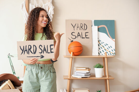 Young African-American woman holding cardboard with text YARD SALE in room of unwanted stuff