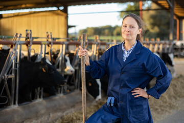 Portrait of positive woman farmer standing in cowshed, holding handle of working tool and smiling.