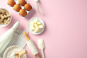 Baking ingredients and tools on a pink background: eggs, butter, milk, flour, sugar cubes, whisk, rolling pin, and spatula. Flat lay, top view.