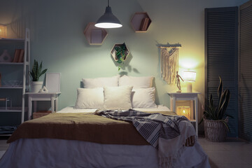 Bed, shelving unit, bedside tables, houseplants and glowing lamps in interior of bedroom