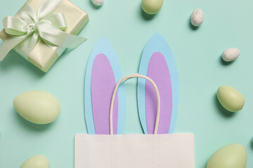 Shopping bag with paper bunny ears, Easter eggs and gift box on turquoise background