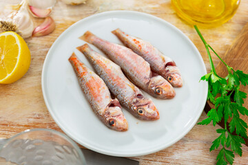 On wooden table there is plate with raw Atlantic fish striped mullus barbatus prepared for frying and baking, and necessary spices and ingredients. Process of cutting fish