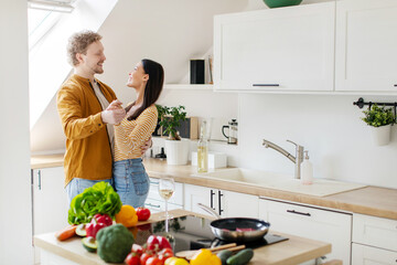 Romantic young couple having fun and dancing while cooking together at kitchen, happy husband hugging his beautiful wife, free space