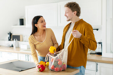 Smiling European couple accurately putting fresh vegetables out of string bag, unpacking groceries at home in kitchen, copy space