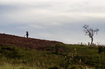 Alone rural worker throwing water on a planting plot on the side of a hill surrounded by native grass with a background of blue sky with rain clouds at dusk
