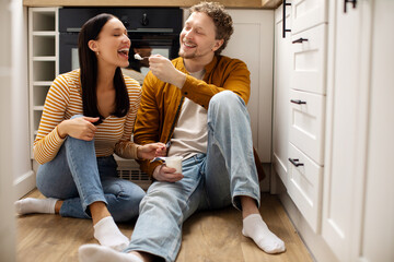 Happy loving couple enjoying time together, man feeding wife with yogurt while sitting on floor in...
