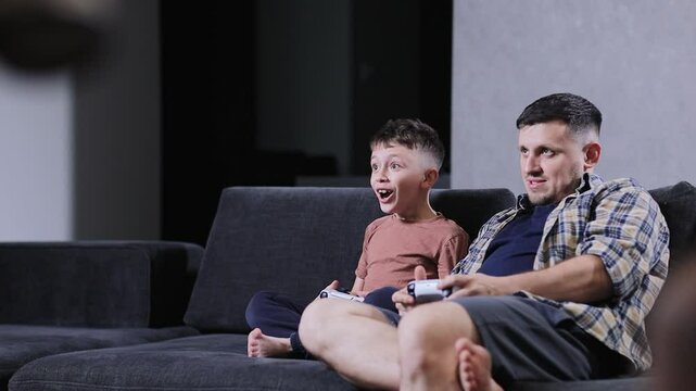 Father and his little son sit on sofa in living room playing video games together at home.