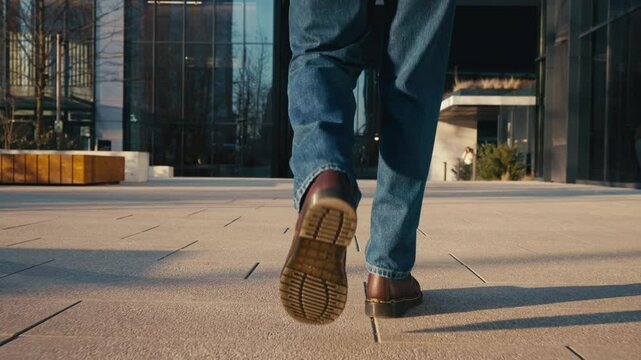 Back view following male feet steps walking street on sunny autumn day. Man wearing blue jeans and leather boots goes urban sidewalk in city. Low angle people legs stepping backlit warm sunlight