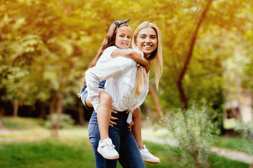 Fototapeta premium Joyful Mom Carrying Little Daughter Piggyback Having Fun Playing In Park Outdoors. Happy Motherhood Concept