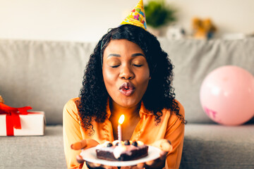 African American Woman Making Birthday Wish And Blowing Candle On Cake Wearing Festive Hat Sitting...