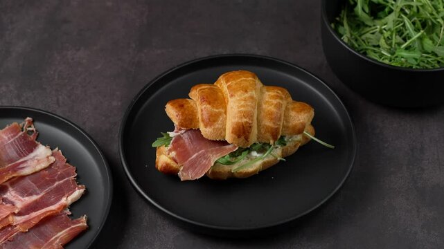 Croissant sandwich with serrano ham and arugula leaves on a black plate