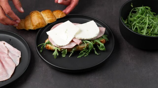 Croissant sandwich with black forest ham and arugula leaves, hands placing slices of panela cheese, healthy food