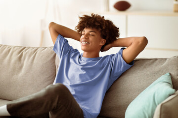 Cheerful black teenager relaxing on comfy couch at home during covid quarantine. Peaceful African...