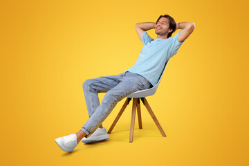 Vertical Portrait Of Happy Casual Millennial Man Relaxing Sitting In Chair Holding Hands Behind Head Leaning Back, Posing With Eyes Closed On Yellow Orange Studio Wall Background, Enjoying Silence