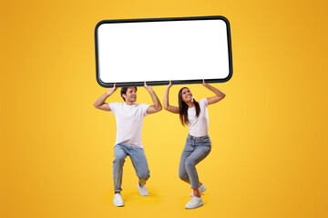 Millennial couple holding huge heavy smartphone with white empty screen on yellow orange studio...