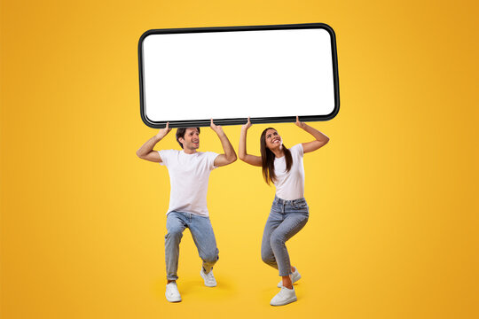 Millennial couple holding huge heavy smartphone with white empty screen on yellow orange studio background. Mock up for mobile app or new website advertising, great offer. Cellphone display template
