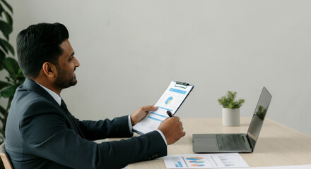 A man in a suit is sitting at a desk with a laptop and a clipboard