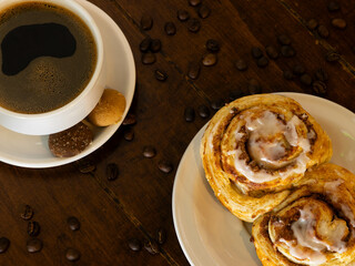 cup of coffee placed on a white plate with a bread on another plate, there are coffee beans scattered on the table