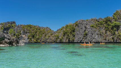 Obraz premium Canoes with tourists sail along the turquoise lagoon. Green vegetation on the steep slopes of coastal karst cliffs. Clear blue sky. Philippines. Palawan. Big Lagoon. Bacuit bay