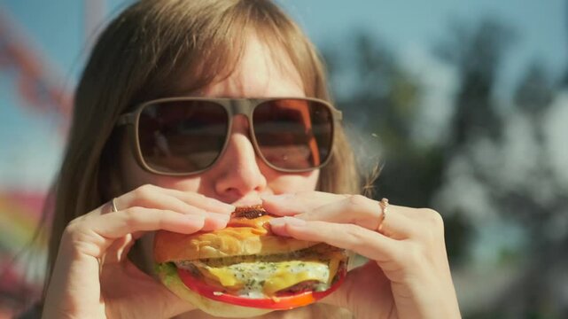 Close up of a brunette woman in sunglasses eagerly bites into a burger on a warm summer day, featuring crispy lettuce, juicy tomato, melted cheese, and a golden, toasted bun.