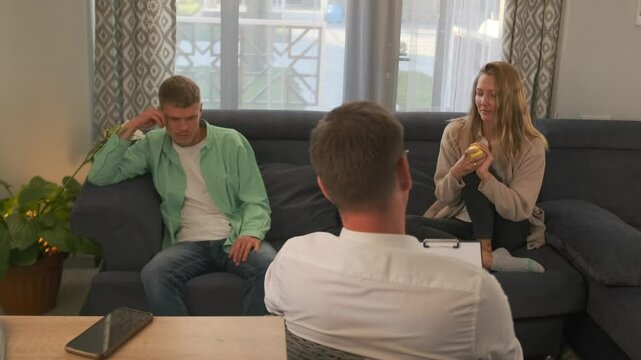 During the session, the woman holding a ball shares her thoughts as the man opposite responds with disagreeing gestures. The psychotherapist observes, analyzing the couple interaction dynamics.
