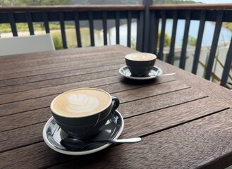 Two cups of coffee on saucers on a wooden outdoor table with a balustrade in background.