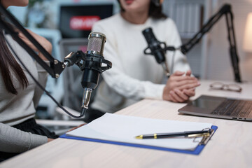 Two individuals are actively engaging in a podcast discussion, using professional microphones and a laptop. A notepad and pen are ready for taking notes on the desk